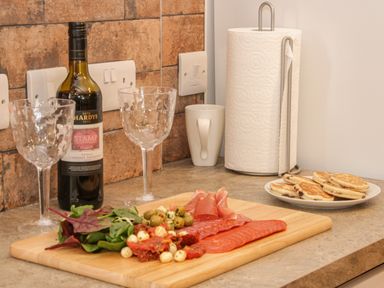 A kitchen countertop with two wine glasses a bottle of wine a paper towel holder a mug a plate with pancakes and a cutting board with greens olives cheese and sliced meats at Conquer Hall in Hendomen near Montgomery