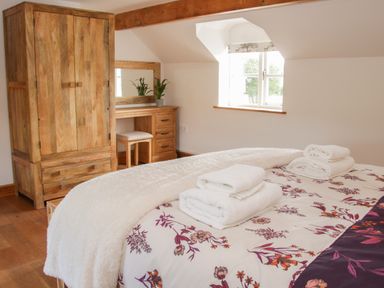 A bedroom with a wooden wardrobe and dressing table with a mirror and stool beside a window with floral blinds and a bed with floral bedding and folded towels at Conquer Hall in Hendomen near Montgomery
