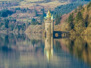 A stone tower with a bridge over a calm lake surrounded by forested hills at Conquer Hall in Hendomen near Montgomery