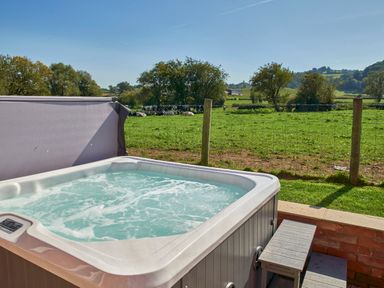 An outdoor hot tub with bubbling water next to a small bench overlooking a fenced green field with cows and trees at Conquer Hall in Hendomen near Montgomery