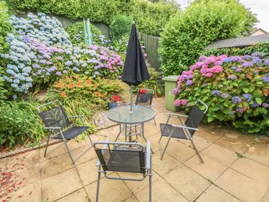 A patio with a glass table and four black chairs surrounded by flowering bushes at 2 Primrose Cottage in Saundersfoot