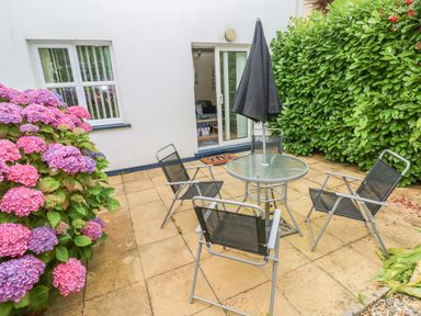 A patio with glass table and four chairs next to a sliding door and surrounded by green hedges and pink flowers at 2 Primrose Cottage Saundersfoot