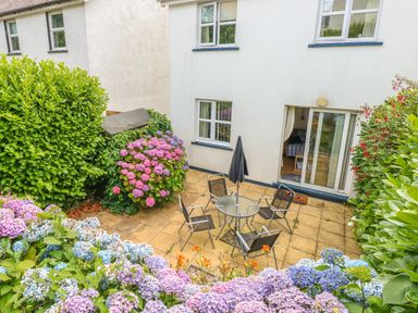 A patio with a glass table and four chairs surrounded by flowering bushes outside a white house with sliding doors at 2 Primrose Cottage in Saundersfoot