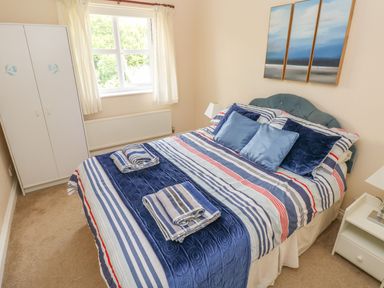 A bedroom with a double bed with striped bedding and pillows a wardrobe a bedside table with a lamp and a three-panel painting above the bed at 2 Primrose Cottage in Saundersfoot