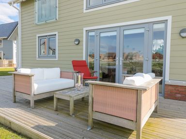 An outdoor wooden deck with two cushioned sofas a red cushioned chair and a wooden coffee table at Waterside in Beadnell