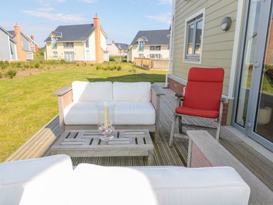 An outdoor wooden deck with white cushioned seating a red cushioned chair and a glass candle holder at Waterside in Beadnell