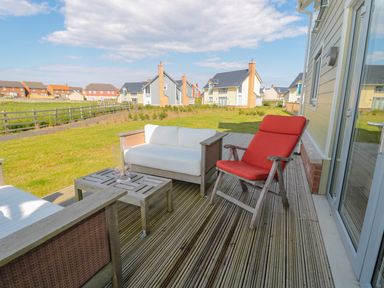 An outdoor patio with wooden chairs and a small table on a deck overlooking a grassy backyard and houses at Waterside in Beadnell