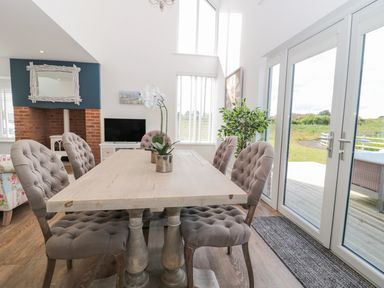 A dining area with a wooden table and upholstered chairs next to large windows overlooking a garden at Waterside in Beadnell