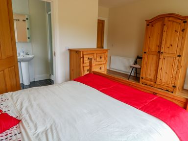 A bedroom with a bed covered with a white and red blanket a wooden dresser a wardrobe a chair and a door leading to a bathroom at Croghan in Portsalon