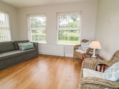 A sitting room with a sofa two wicker chairs a small wooden table with a lamp and windows with vertical blinds at Croghan in Portsalon
