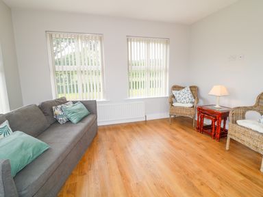 A room with a gray sofa with cushions two wicker chairs a small wooden table with a lamp and windows with vertical blinds at Croghan in Portsalon
