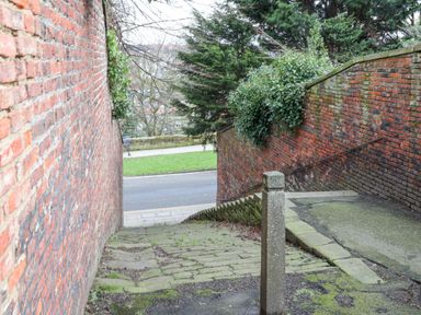 A narrow outdoor stone staircase between two brick walls with greenery hanging over one wall at 20A St Hildas in Whitby