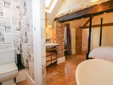 A bedroom with a four-poster bed a freestanding bathtub wooden flooring and an adjacent toilet with newspaper-themed wallpaper at The Hemmel in Faringdon