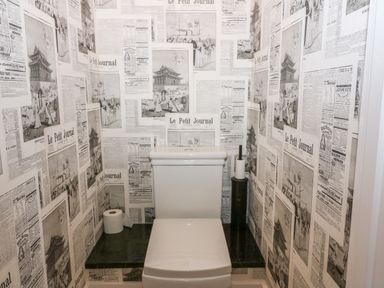 A small bathroom with a toilet and black shelf with a toilet paper roll and brush surrounded by newspaper print wallpaper at The Hemmel in Faringdon