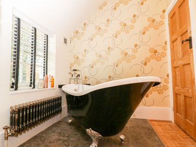 A bathroom with a black clawfoot bathtub floral wallpaper a wooden door a radiator and a window with blinds at The Hemmel in Faringdon