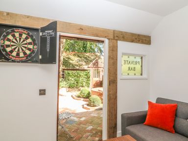 A dartboard on the wall a gray sofa with an orange pillow a window with private bar sign and a door opening to a brick patio with plants at The Hemmel in Faringdon