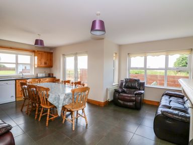 A dining area with wooden chairs around a table and a kitchen with wooden cabinets next to a sitting area with black leather armchair and sofa at Doornogue in Churchtown near Fethard-on-Sea