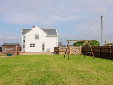 A white house with a gray roof next to a wooden shed and a green yard with a swing set at Doornogue in Churchtown near Fethard-on-Sea