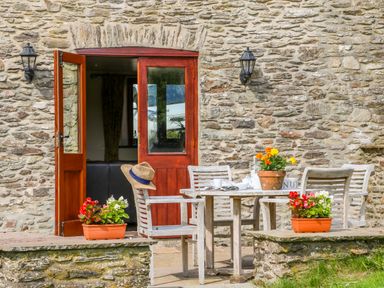 An outdoor stone patio with wooden table and chairs next to a stone wall with open wooden door and potted flowers at Great Bradley Cottage in Withypool