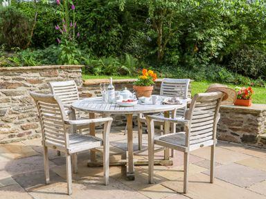 An outdoor patio with a round wooden table and four wooden chairs surrounded by stone walls and greenery at Great Bradley Cottage in Withypool