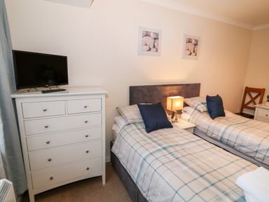 A bedroom with two single beds with checked bedding a white chest of drawers with a television and a wooden chair at 4 Ty Rhosyn in Morfa Bychan