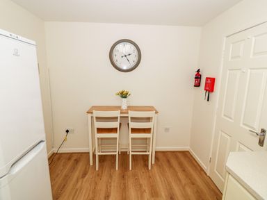 A small dining table with two chairs a clock on the wall a fire extinguisher and a fire blanket in a kitchen at 4 Ty Rhosyn in Morfa Bychan