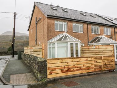 A corner house with a conservatory and wooden fence on a street with hills in the background at 4 Ty Rhosyn in Morfa Bychan