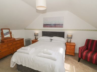 A bedroom with a double bed side tables lamps a wooden dresser a mirror and a striped armchair at 4 Ty Rhosyn in Morfa Bychan