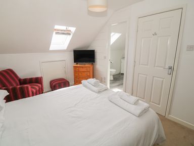 A bedroom with a bed, white towels, a TV on a wooden dresser, a red striped armchair, and an open door showing a bathroom at 4 Ty Rhosyn in Morfa Bychan