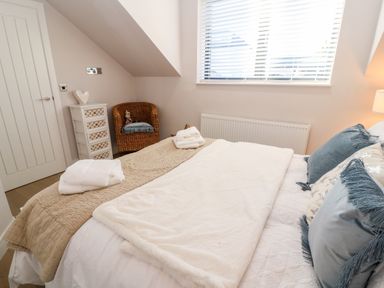 A bedroom with a bed with pillows and blankets a wicker chair a small white drawer unit and a window with blinds at Driftwood Haven in Carbis Bay