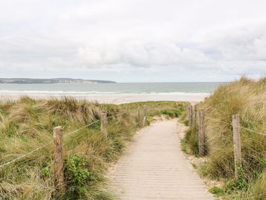 A wooden boardwalk path leading through sand dunes to a beach with the ocean and distant hills visible at Driftwood Haven in Carbis Bay