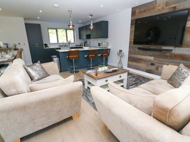 An open living room and kitchen with beige sofas a wooden coffee table a mounted television and a kitchen island with stools at Driftwood Haven in Carbis Bay