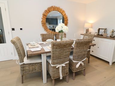 A dining room with a wooden table and wicker chairs a round wooden framed mirror on the wall a sideboard with a lamp clock and decor at Driftwood Haven in Carbis Bay