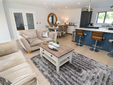 An open living area with beige sofas a center table on a shaggy rug a dining table with wicker chairs and a kitchen island with bar stools at Driftwood Haven in Carbis Bay