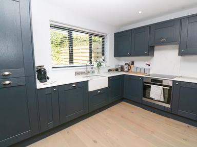 A kitchen with blue cabinets, a white sink under a window, a built-in oven, and wooden flooring at Driftwood Haven in Carbis Bay