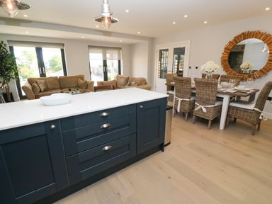 An open living and dining area with a kitchen island brown sofas a dining table with wicker chairs and a round wooden mirror on the wall at Driftwood Haven in Carbis Bay