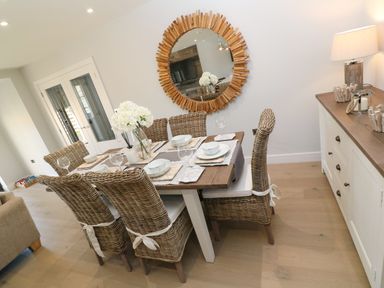 A dining room with a wooden table set with dishes glassware and a vase of flowers with wicker chairs a round wooden framed mirror on the wall and a sideboard with a lamp and decor at Driftwood Haven in Carbis Bay