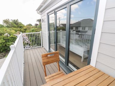 A balcony with a wooden table and chair next to glass sliding doors and white railing surrounded by greenery at Driftwood Haven in Carbis Bay