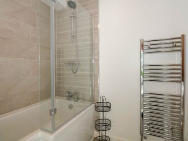 A bathroom with a bathtub shower, glass shower screen, wall-mounted showerhead, and a chrome heated towel rail at Yr Hen Dy Capel in Blaenau Ffestiniog