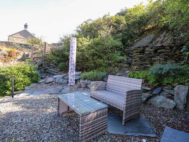 An outdoor seating area with a wicker bench and glass table surrounded by rocks and greenery at Yr Hen Dy Capel in Blaenau Ffestiniog