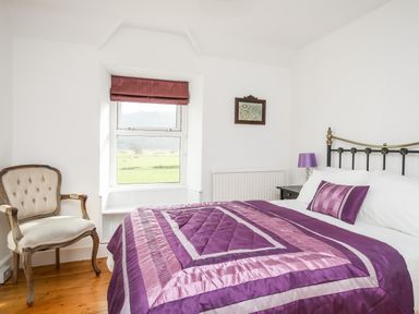 A bedroom with a purple bedspread a chair a window with a red blind and a small framed picture at Glyn Morfa in Harlech