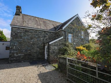 A stone house with a pitched roof and gravel driveway with a garden and metal gate at Glyn Morfa in Harlech