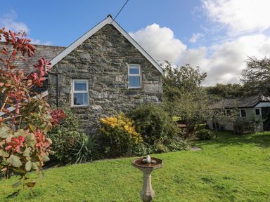 A stone house with two windows and garden with bird bath and trees at Glyn Morfa in Harlech