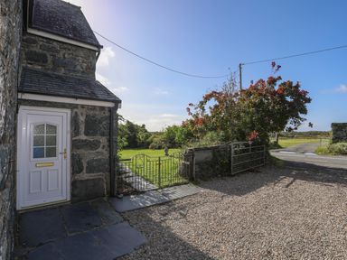A stone house with a white door next to a metal gate and a gravel driveway at Glyn Morfa in Harlech