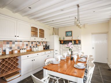 A kitchen with a dining table set with plates and glasses a flower vase on the table white cabinets and a refrigerator Glyn Morfa Harlech