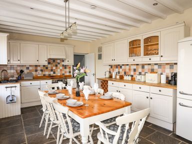 A kitchen with a wooden dining table set with plates bowls glasses and napkins surrounded by white chairs with patterned cushions at Glyn Morfa in Harlech