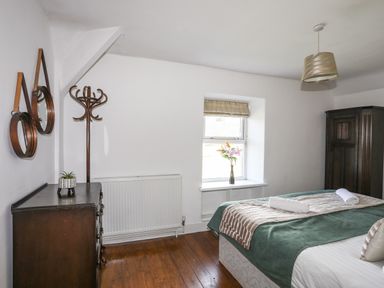 A bedroom with a bed, a window with flowers, a chest of drawers, a coat rack, and two round mirrors on the wall at Glyn Morfa in Harlech