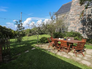 An outdoor patio with wooden table and chairs next to a stone building and grassy lawn at Glyn Morfa in Harlech