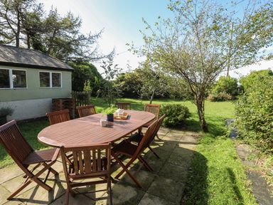 A garden with a wooden table and six chairs on a stone patio near a small building at Glyn Morfa in Harlech
