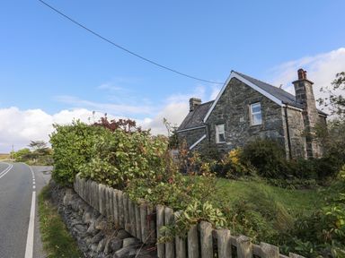 A stone house with a chimney and a garden with bushes and wooden fence next to a road at Glyn Morfa in Harlech